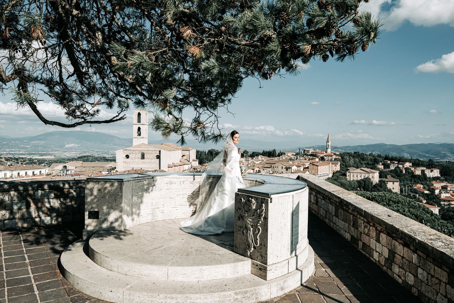 Bride in flowing white dress stands on scenic overlook with panoramic city views.