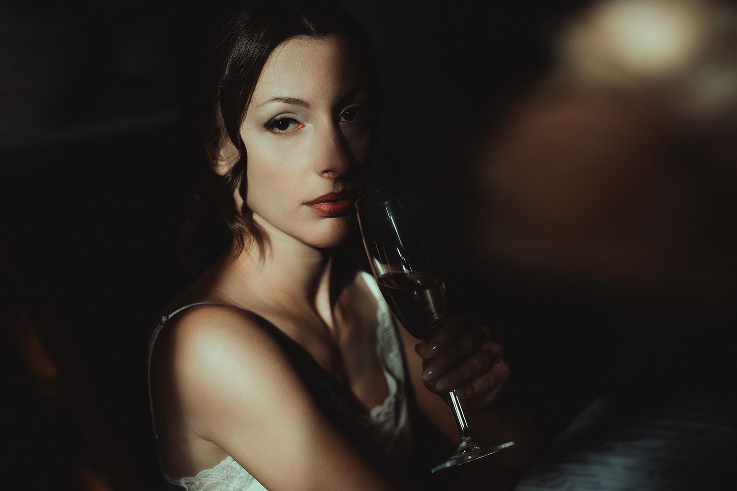 Elegant woman in evening attire holds wine glass in dramatic low-key lighting.