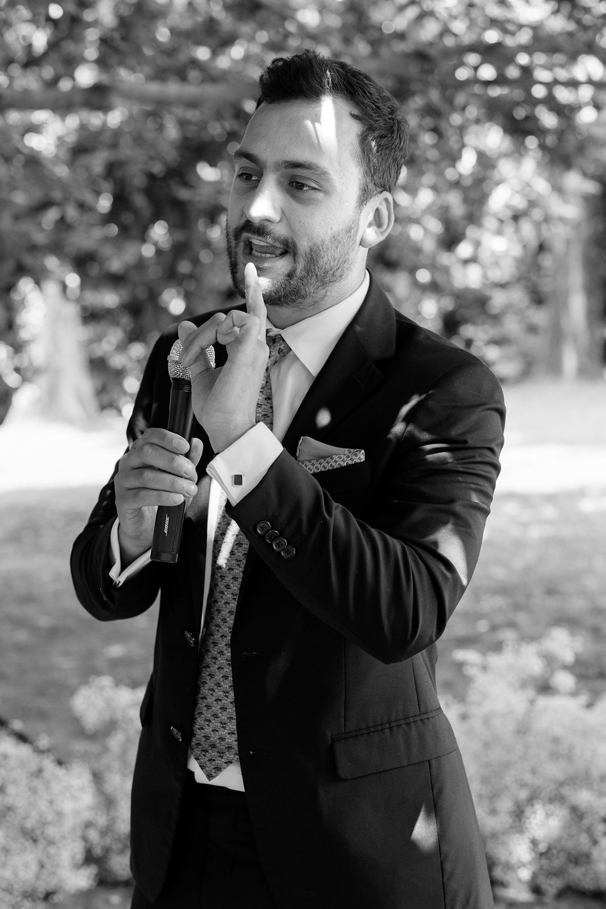 Man in formal suit making expressive gestures while speaking outdoors.