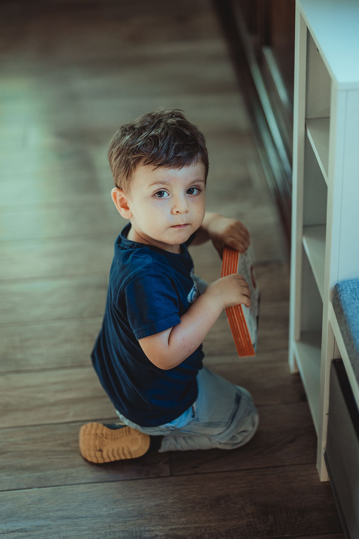 Child in blue shirt crawling and playing under a table on hardwood floor.