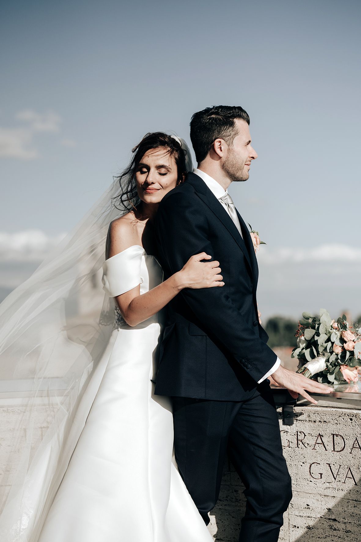 Romantic wedding portrait of couple embracing on stone terrace with flowing veil.