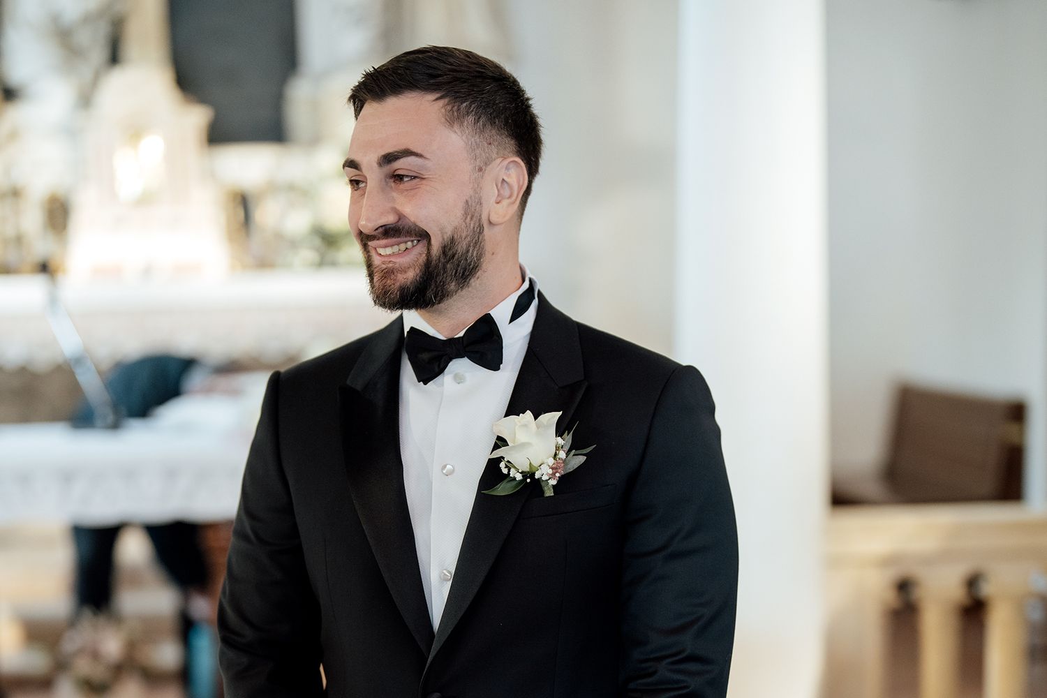 Groom in classic black tuxedo and bowtie smiles during wedding ceremony.