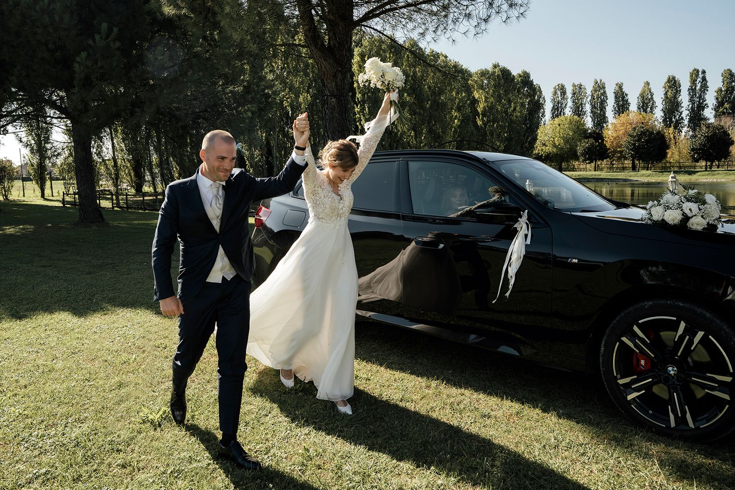 Wedding party poses next to luxury vehicles on a sunny day outdoors.