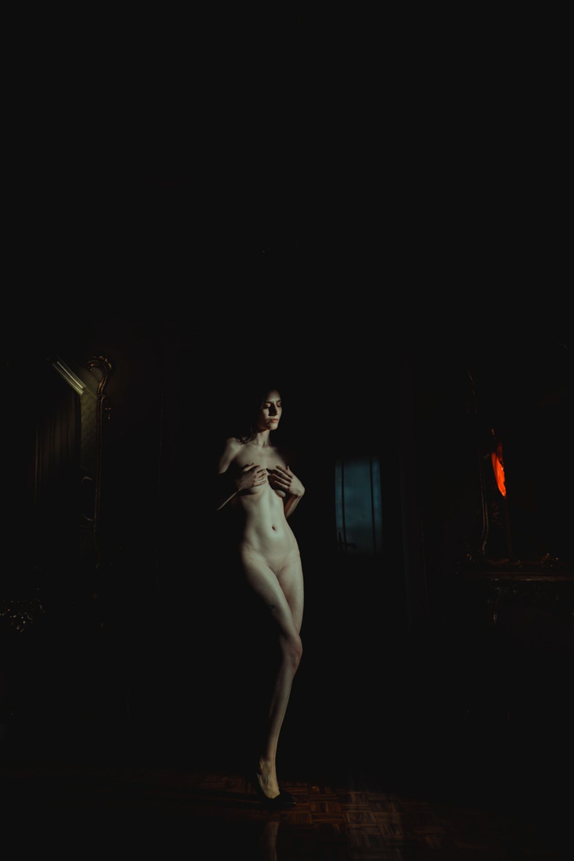 A nude figure stands in dramatic low-key lighting against a dark background with subtle warm tones.