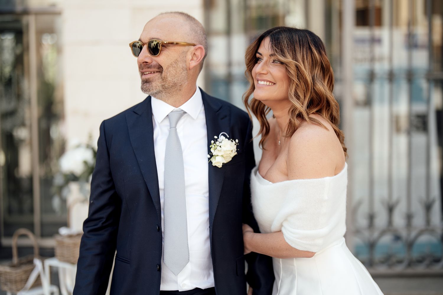 A stylish couple in wedding attire poses together outside a building with elegant architecture.