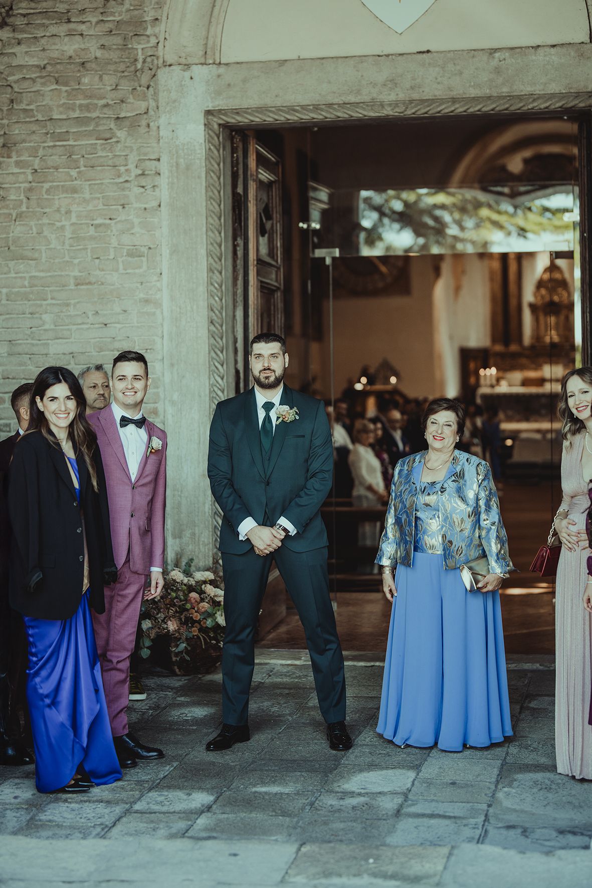 Wedding party poses under stone archway wearing blue and purple formal attire.