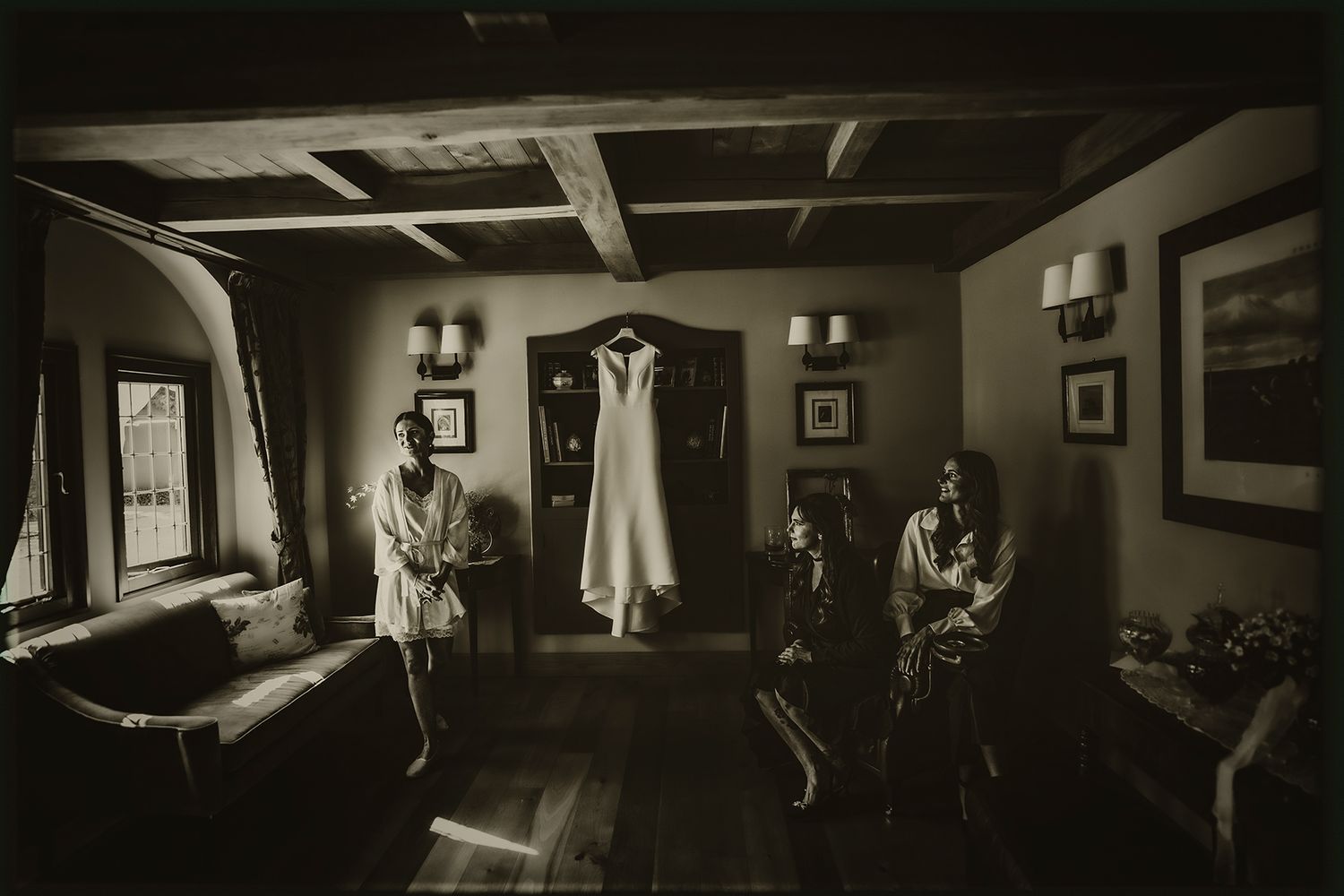 A vintage black and white photograph shows a wedding dress hanging in a rustic room with antique decor and window light.