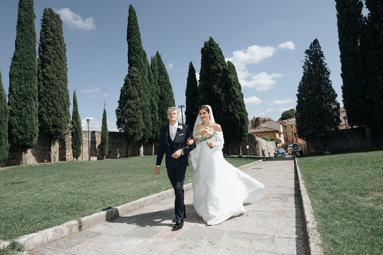 Wedding couple walks along cypress tree-lined path on sunny Italian afternoon.