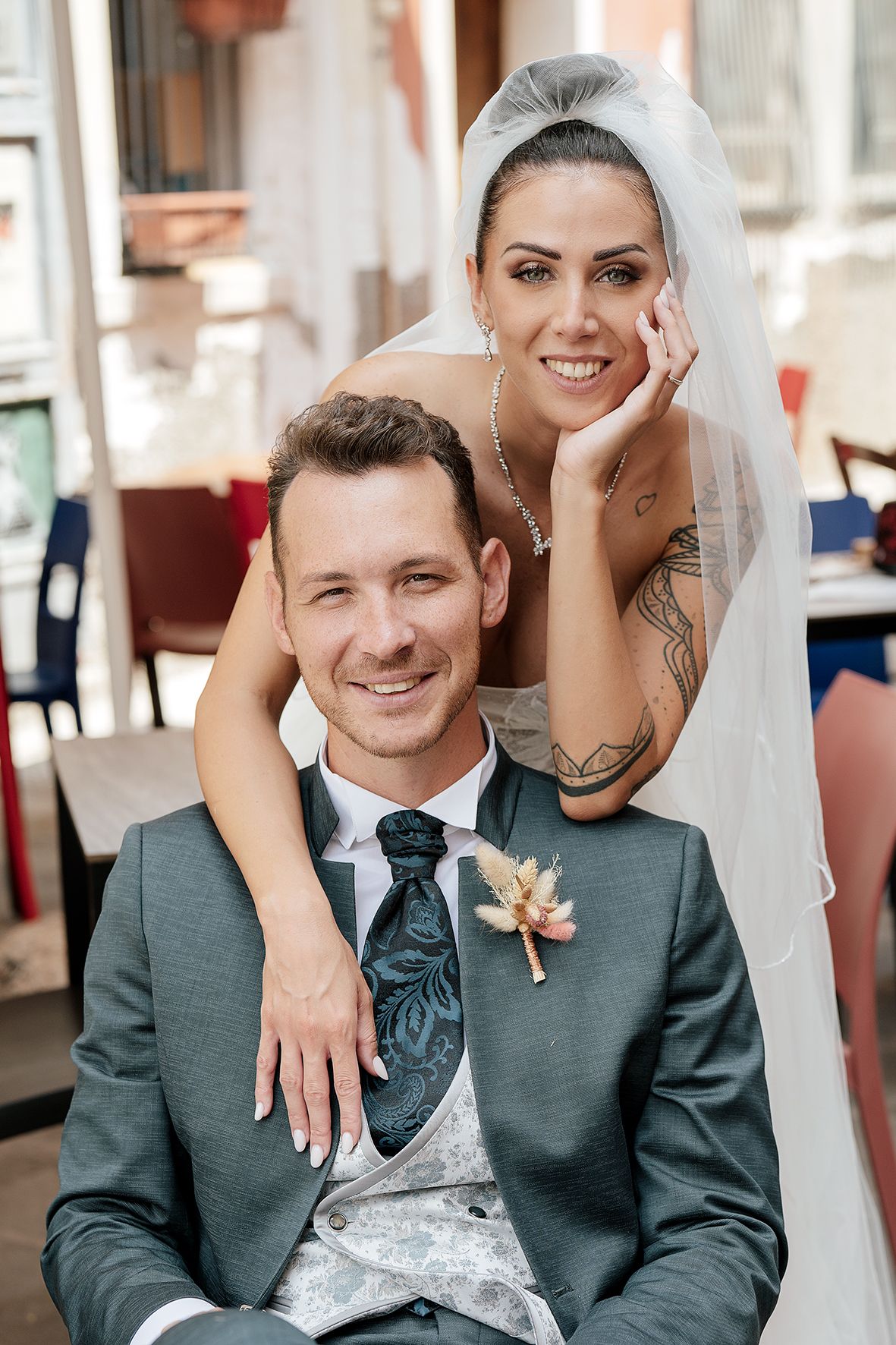 A tender wedding portrait showing the bride in white veil leaning over the groom in a grey suit with boutonniere.
