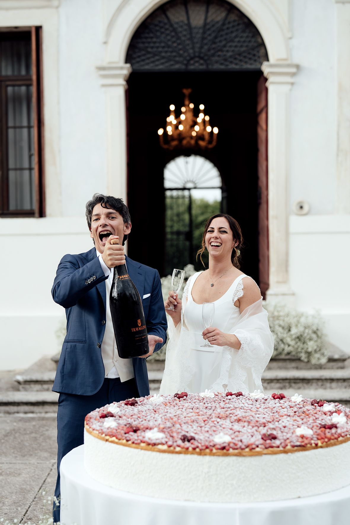 Couple shares a joyful moment while cutting their wedding cake decorated with rose petals.
