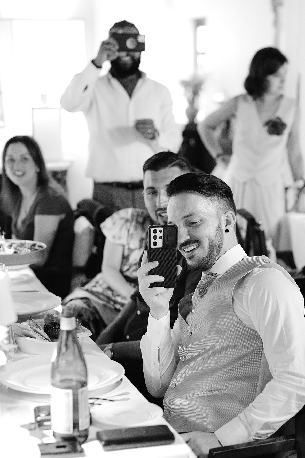 Wedding reception guests sharing laughs at dining tables in black and white photography.