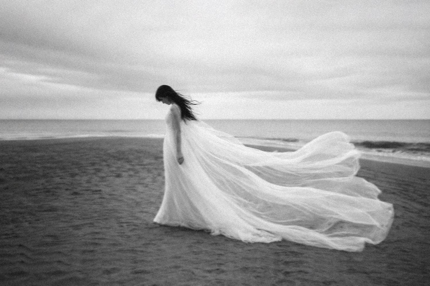An ethereal white gown flows in the coastal breeze as a figure stands on sandy shores.