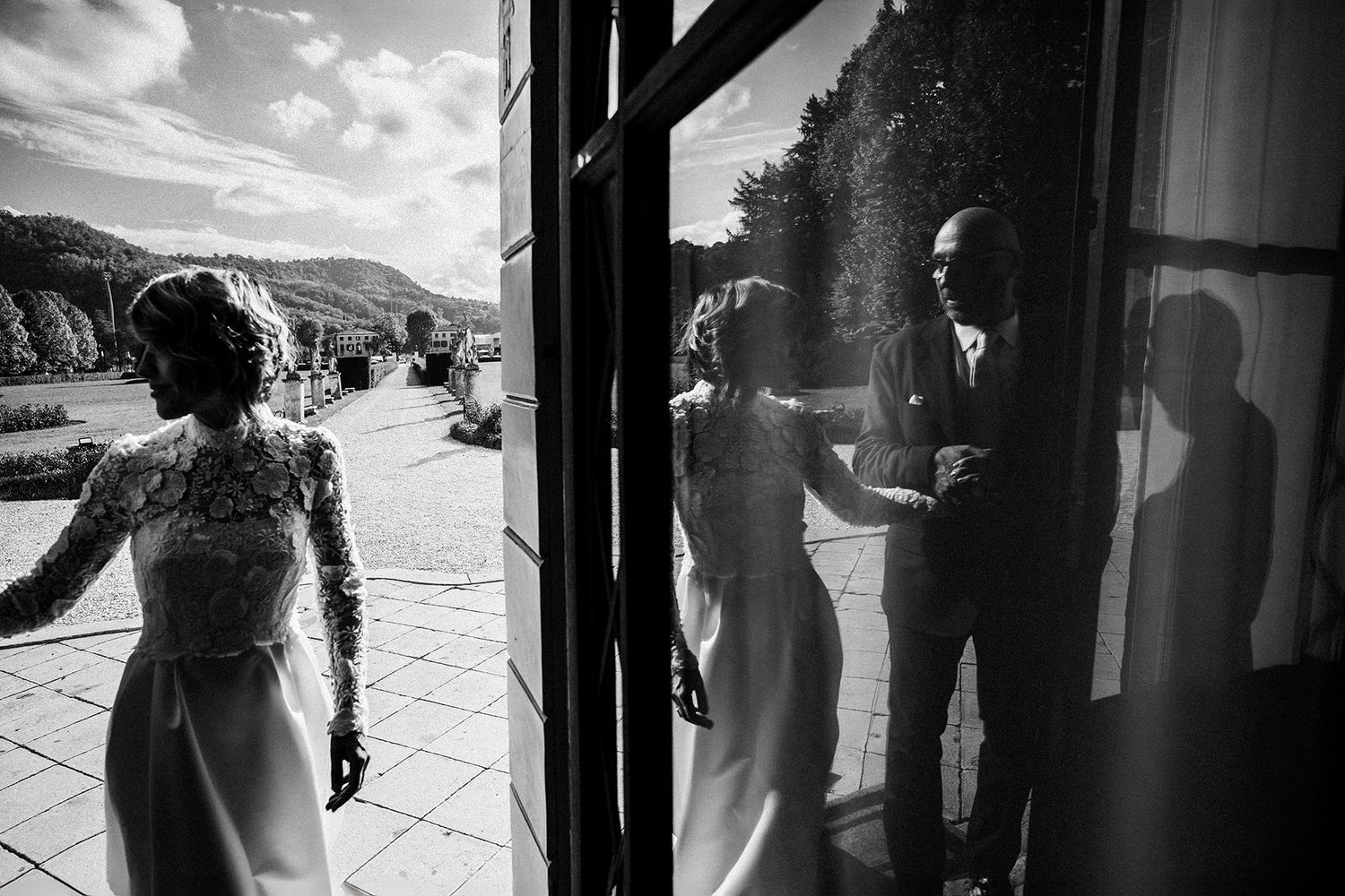 Black and white wedding photo capturing bride in lace gown with reflection of couple in glass door at elegant venue.