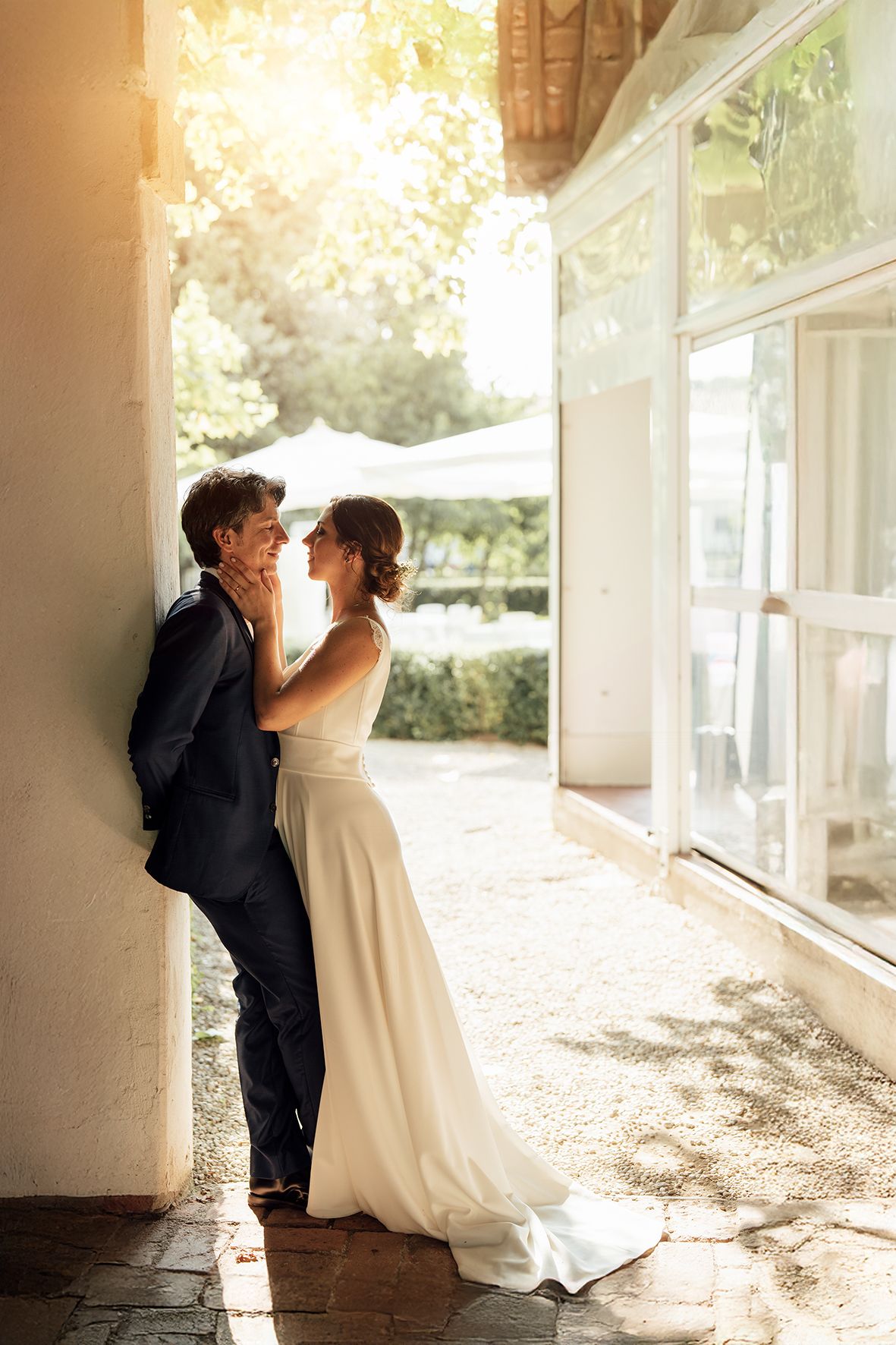 Sunlit wedding portrait of couple in elegant attire sharing an intimate moment by a white wall.