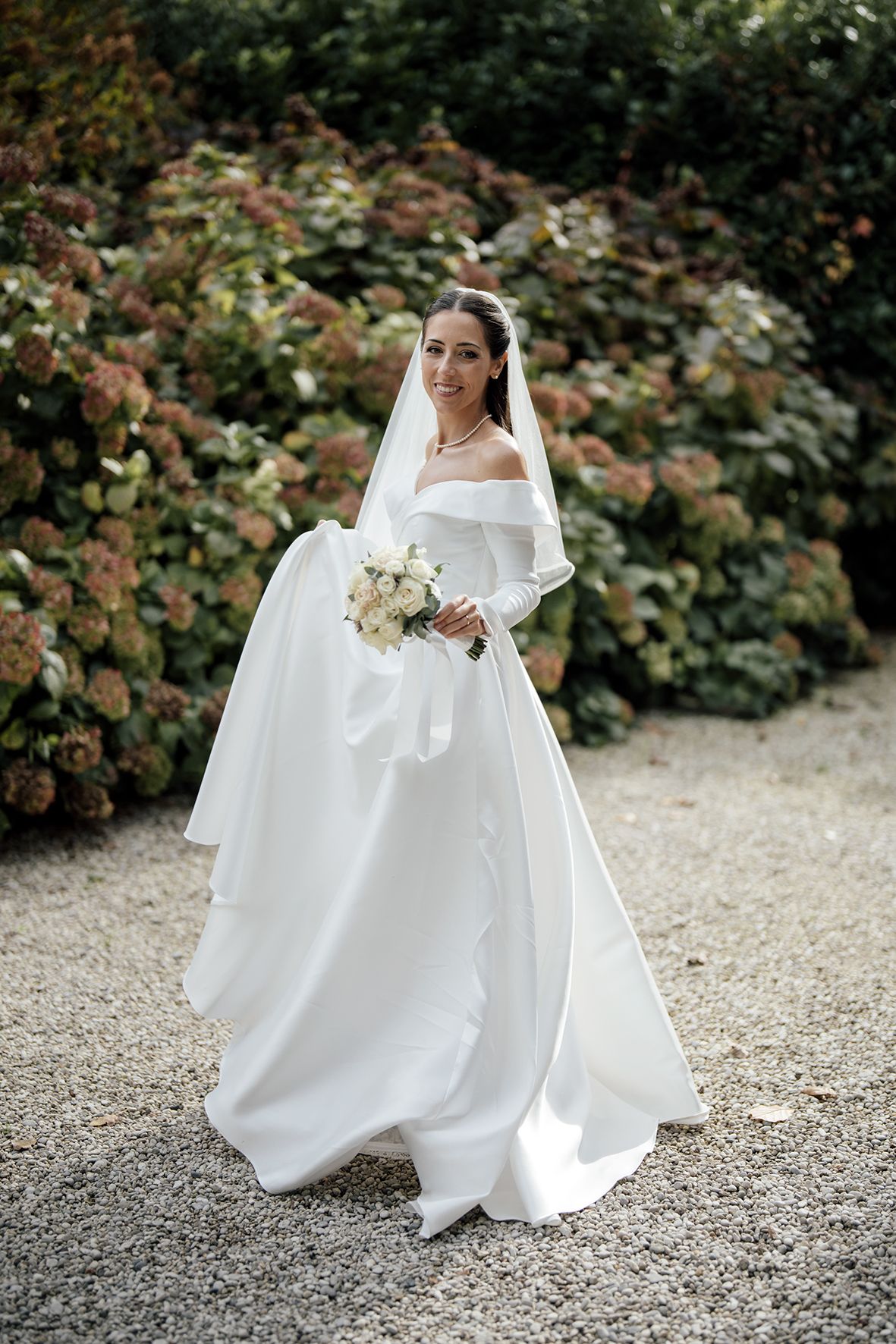 A bride in an elegant off-shoulder ball gown poses in a garden setting with a white bouquet.
