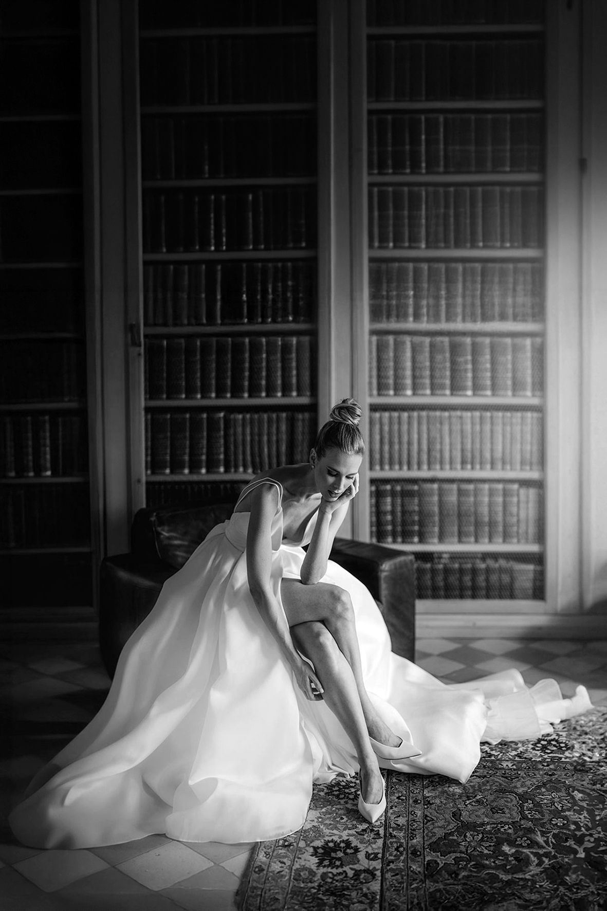 A bride in a flowing white gown adjusts her shoe in an elegant library with floor-to-ceiling bookshelves.