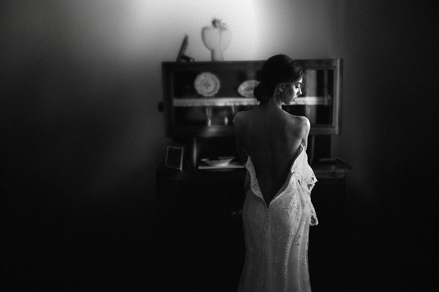 Black and white photo of a bride in a backless wedding gown standing at a wooden dresser in a dimly lit room.