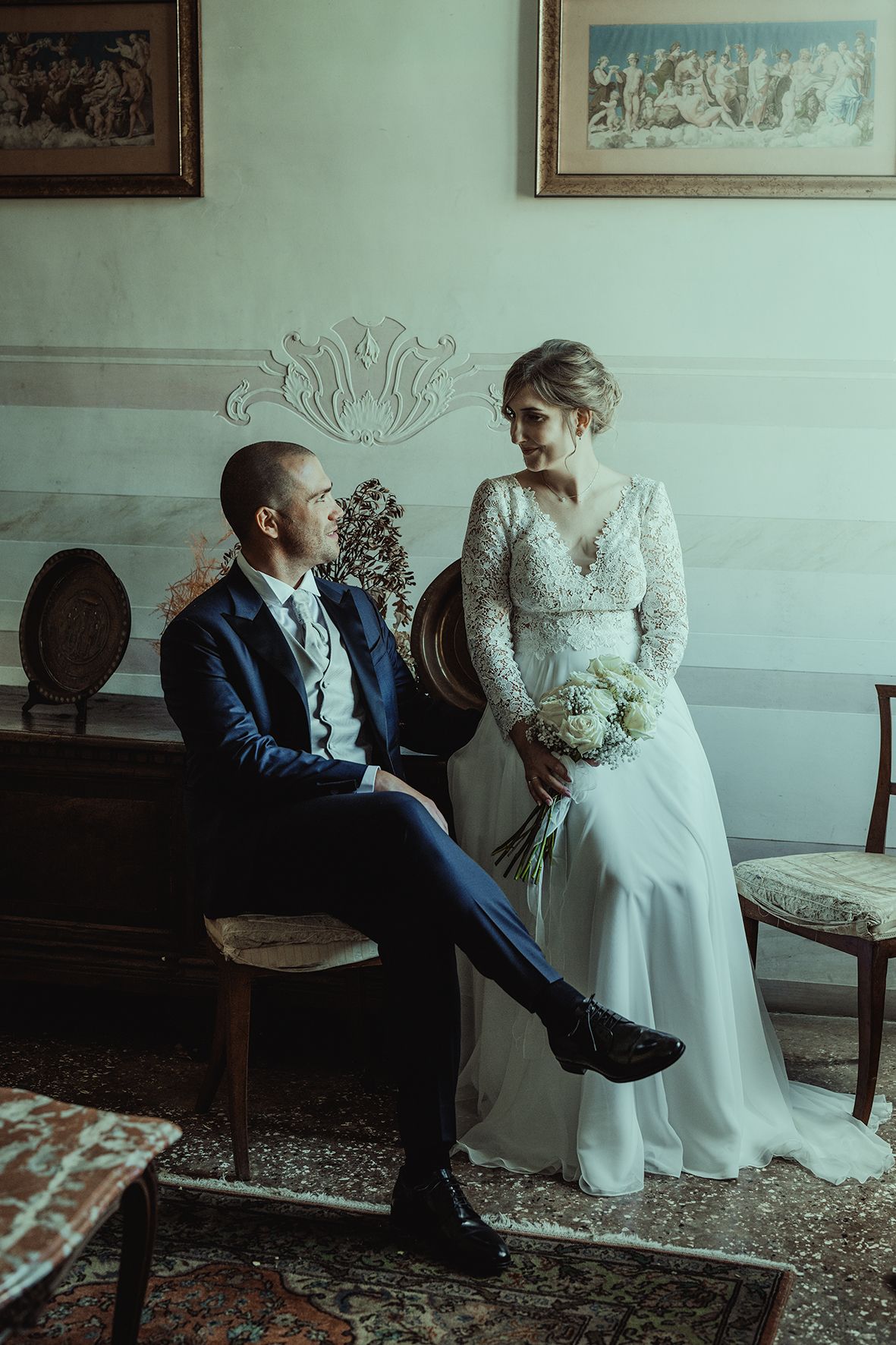 Elegant couple in wedding attire seated in classic interior with ornate wall details.
