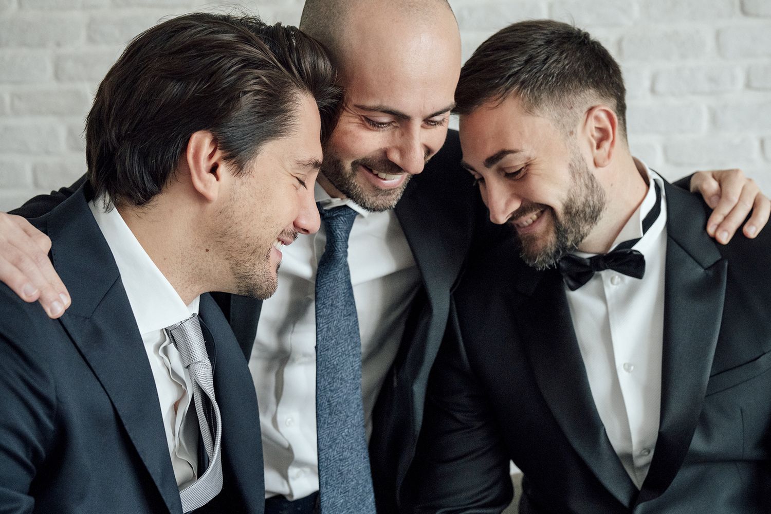 Group of men in suits sharing candid laughs and embraces against white brick wall.