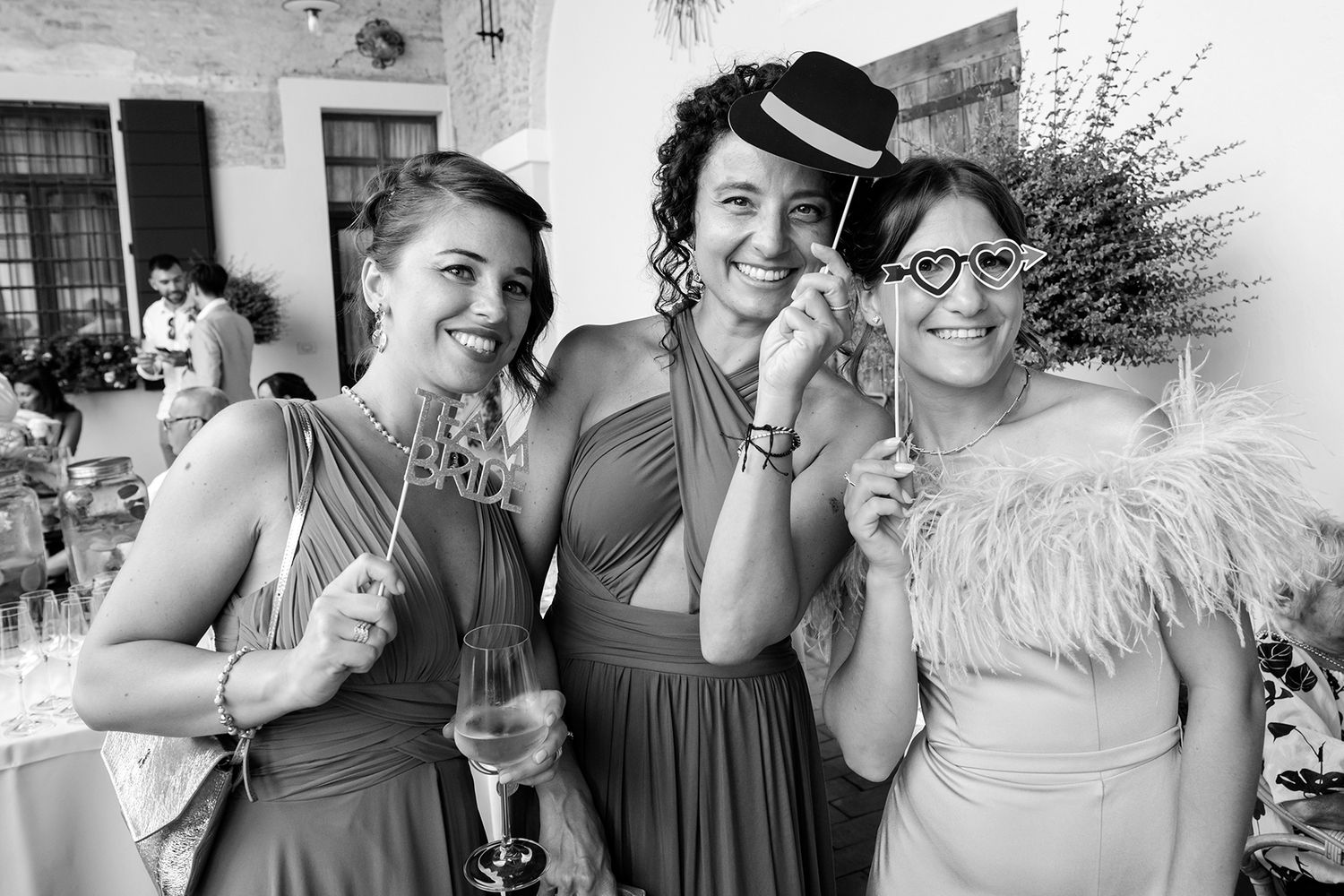 Wedding party members pose with fun props and joyful expressions at outdoor celebration.