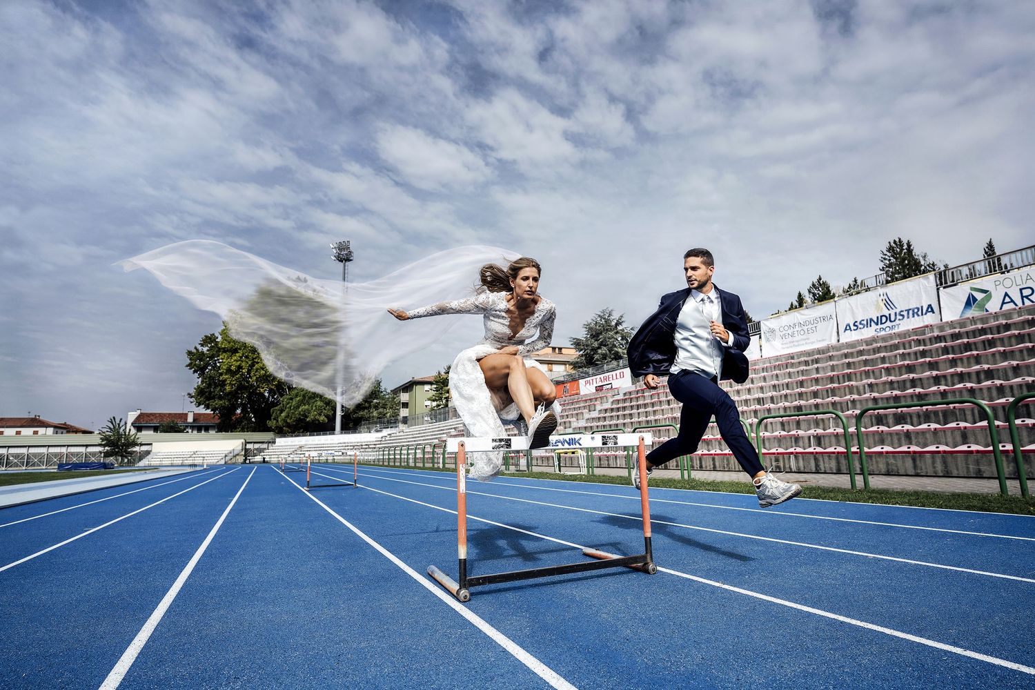 Bride in a flowing veil leaps over a hurdle on a blue running track while the groom in a suit runs beside her.