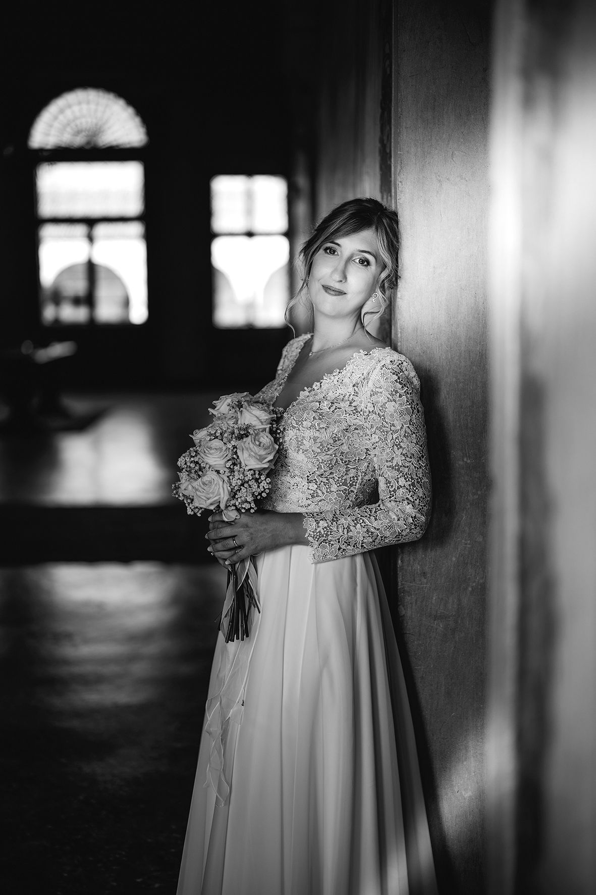 Elegant bride in lace and silk wedding gown poses by window with bouquet in dramatic black and white portrait.