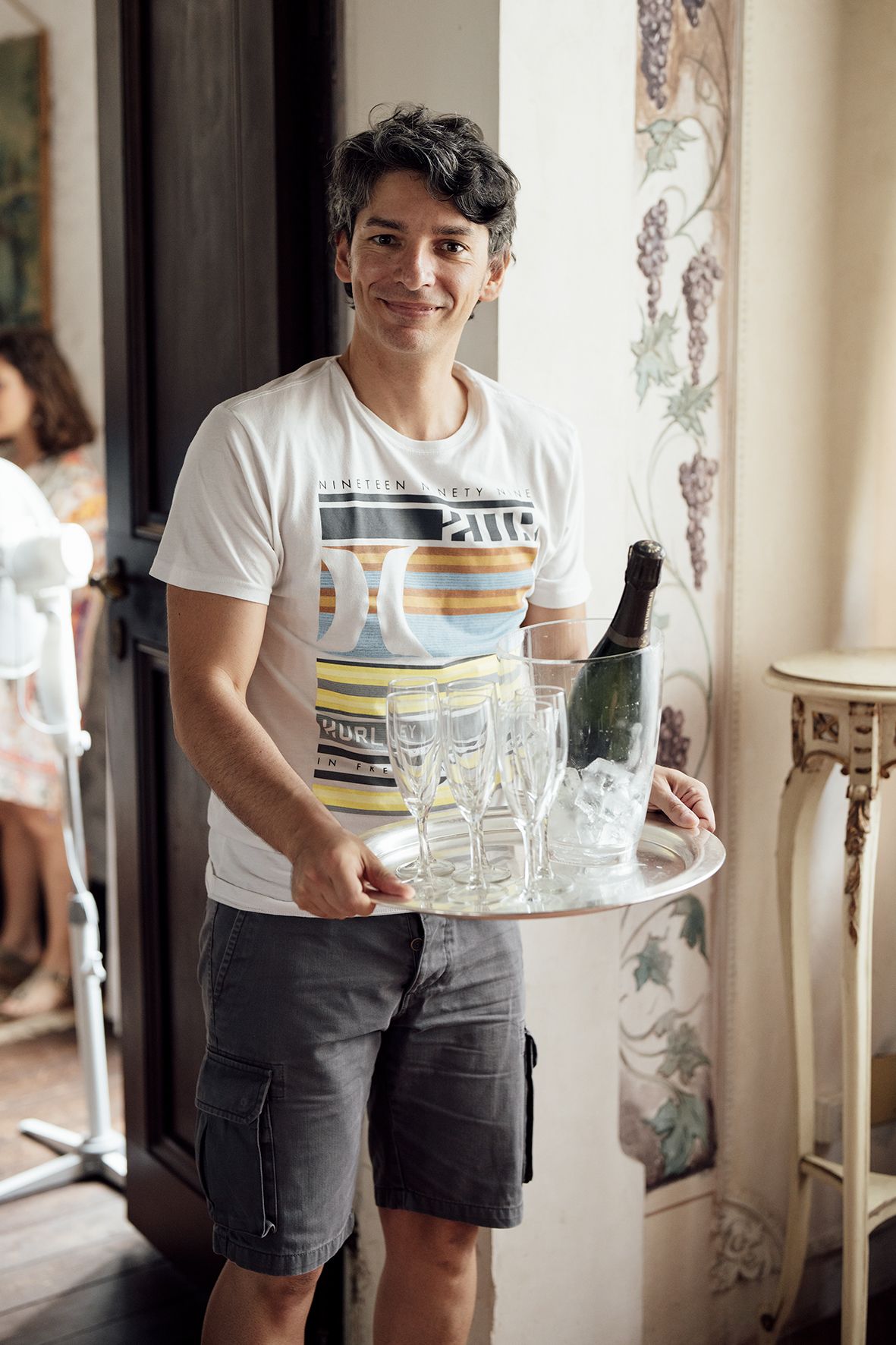 Person in white t-shirt carrying glassware and dishes on a serving tray in an elegant room.