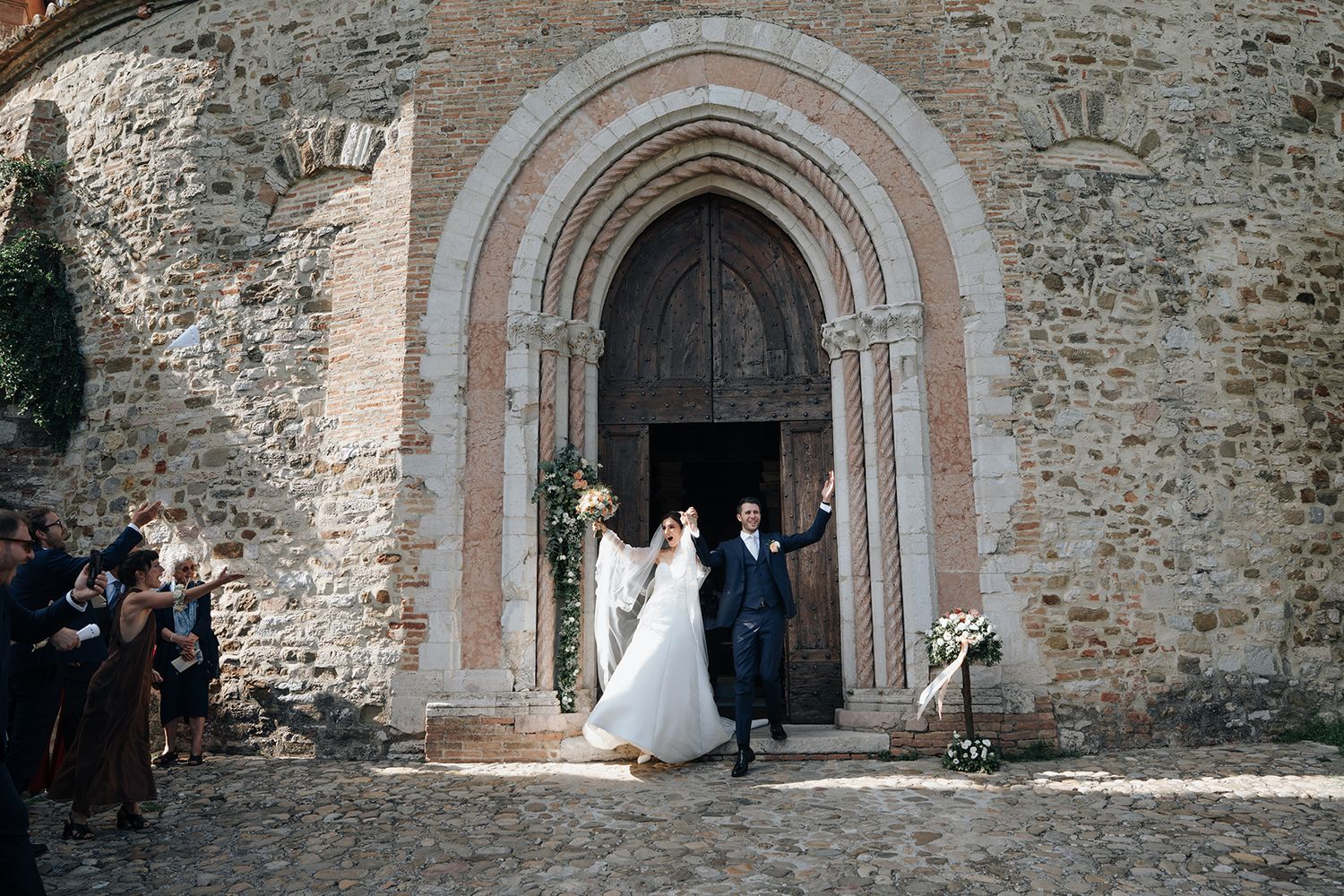 Newlyweds exit through an ancient stone church archway as guests look on.