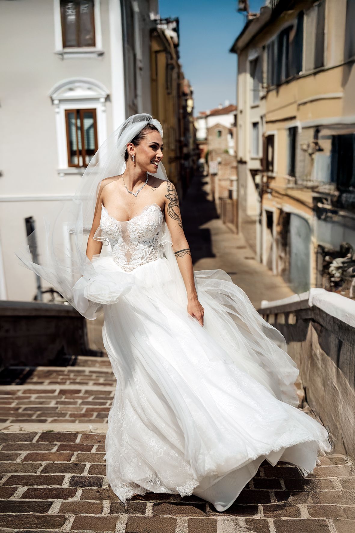 Bride in flowing white dress twirls gracefully on cobblestone street in European town setting.