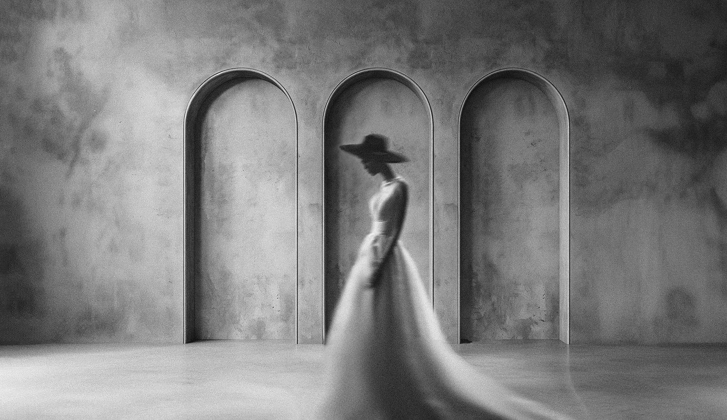 Black and white photo of elegant woman in flowing white gown and wide-brim hat standing before arched stone wall.