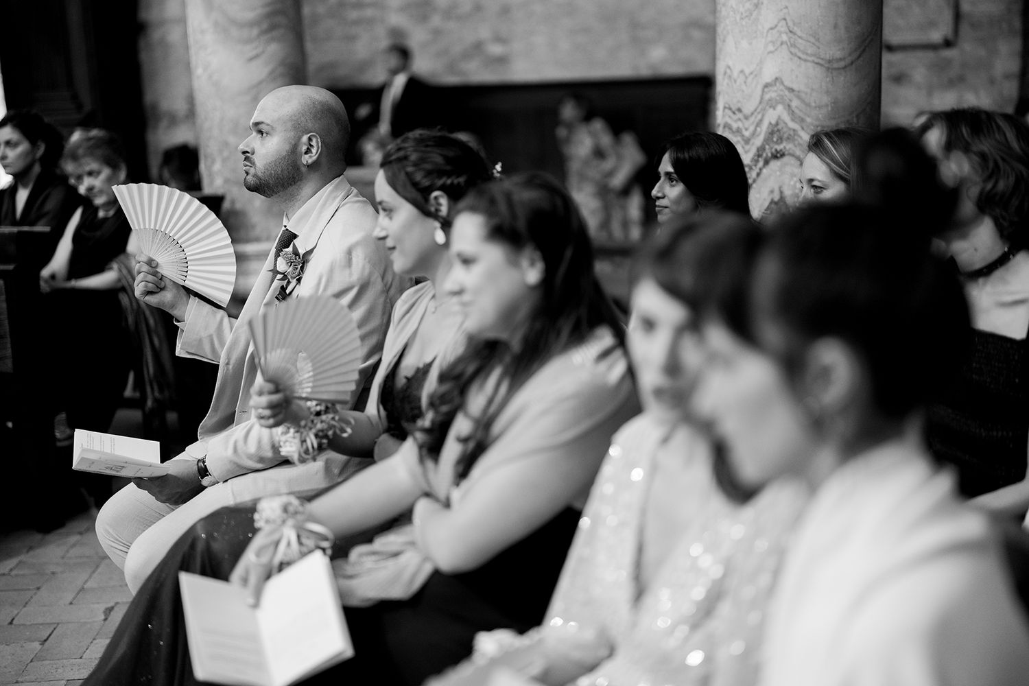Wedding guests seated in church pews with one person holding a decorative fan.