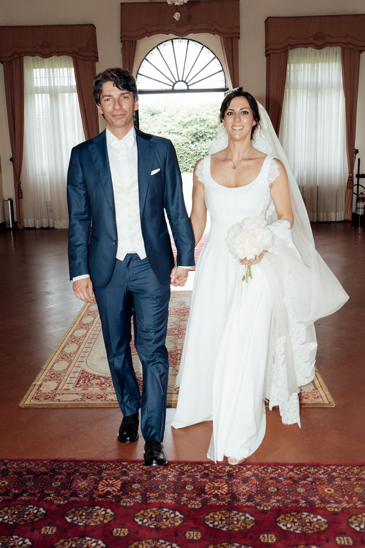 Wedding couple walking through elegant interior with ornate rugs and window treatments.