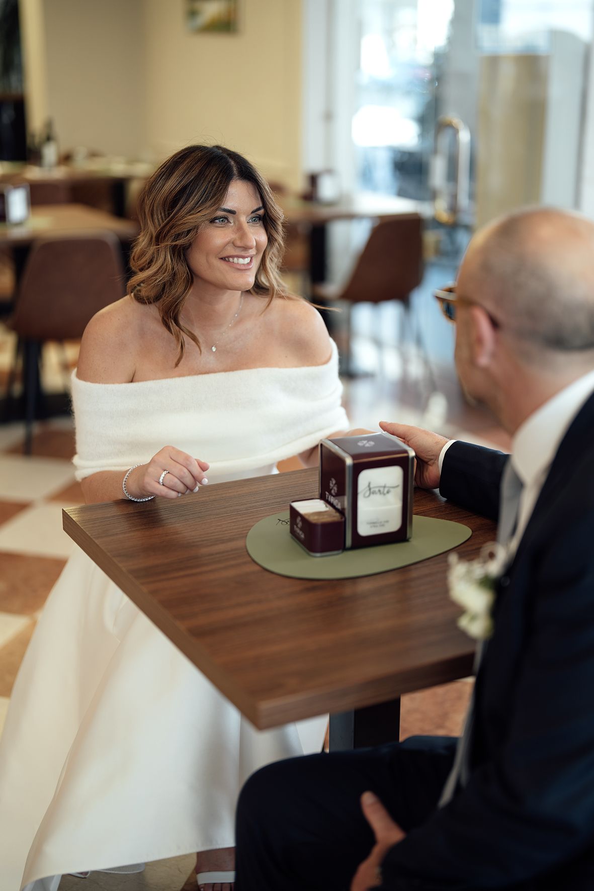 A bride in an off-shoulder dress smiles while seated at a table with a jewelry box.