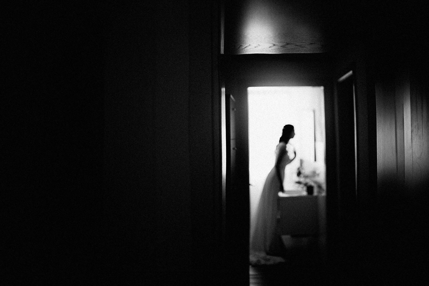 Black and white photo of a bride in a white gown standing in a doorway, backlit by bright light.