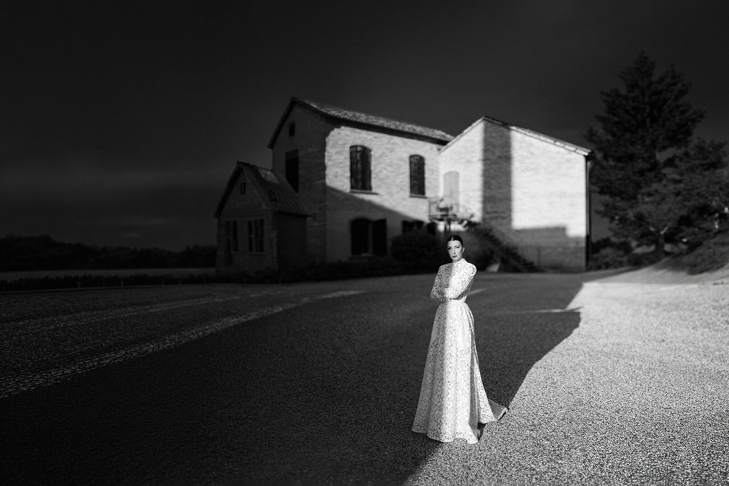 Black and white photo of a woman in a white dress standing on a road beside a historic stone building at dusk.