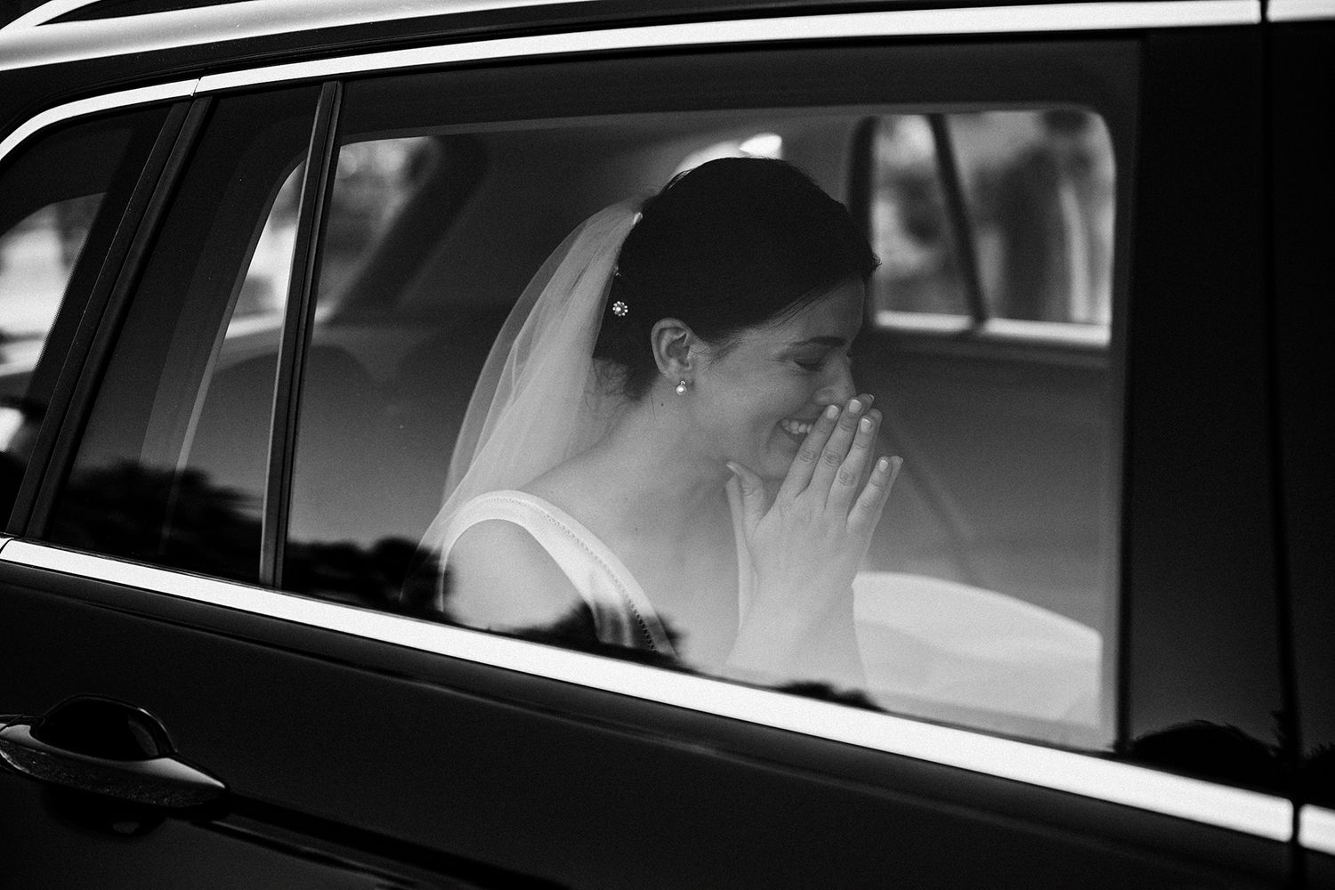 Black and white photo of a bride in a veil sitting in a car, hands clasped near her face on her wedding day.