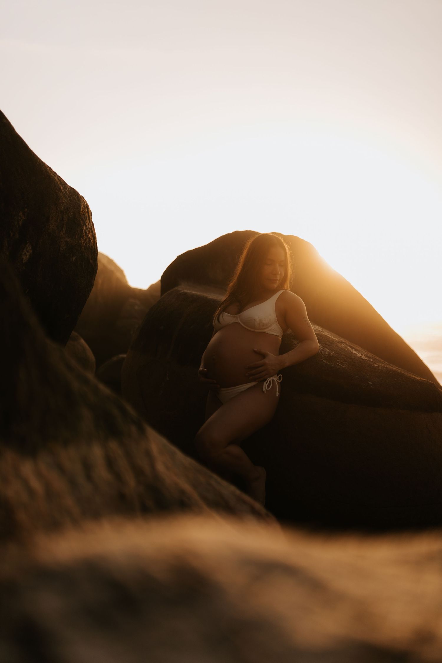 Silhouetted maternity pose against sunset and rocky coastline.