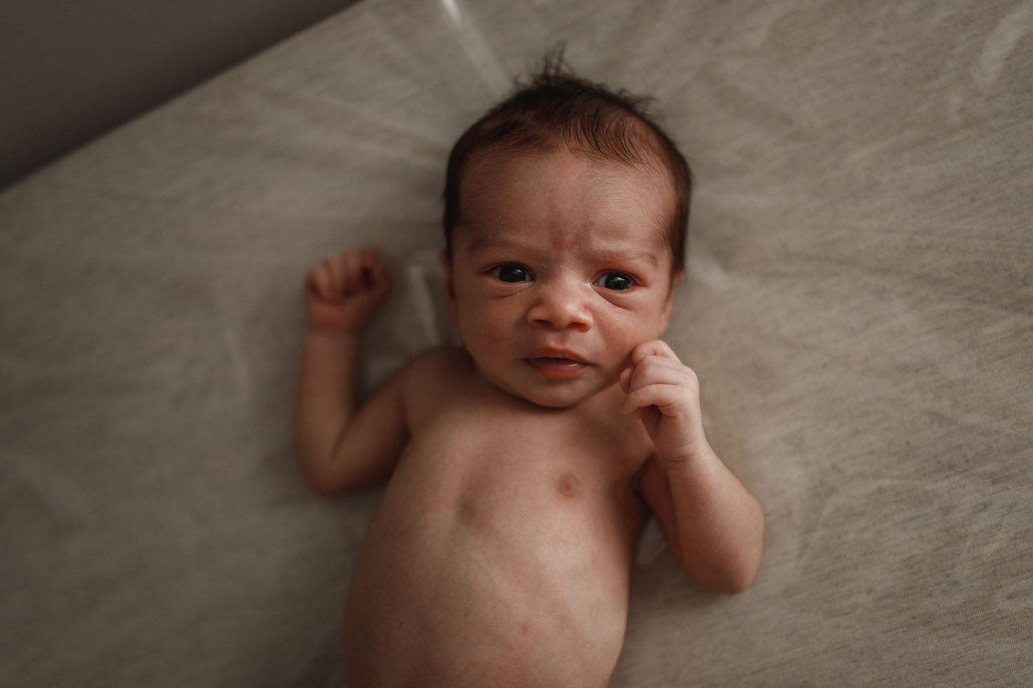 A newborn baby lying on soft gray fabric making expressive faces.