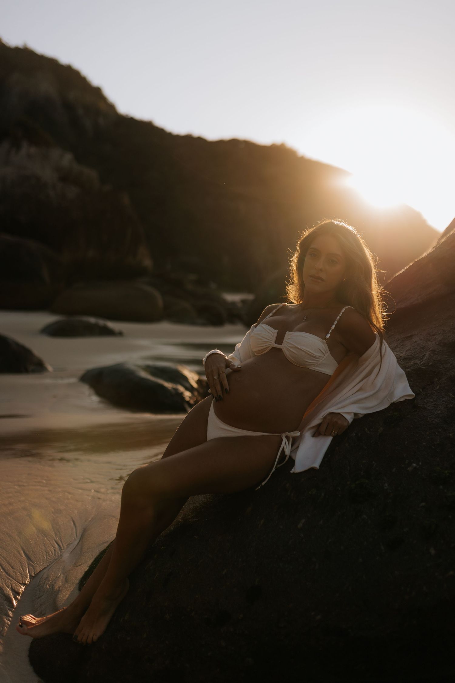 Backlit figure reclining on rocky beach during sunset.