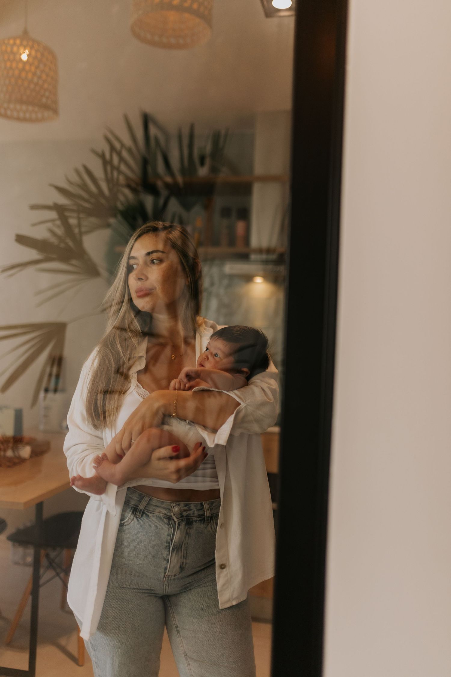 A casual indoor portrait of a parent holding a baby near tropical plants and woven pendant lights.