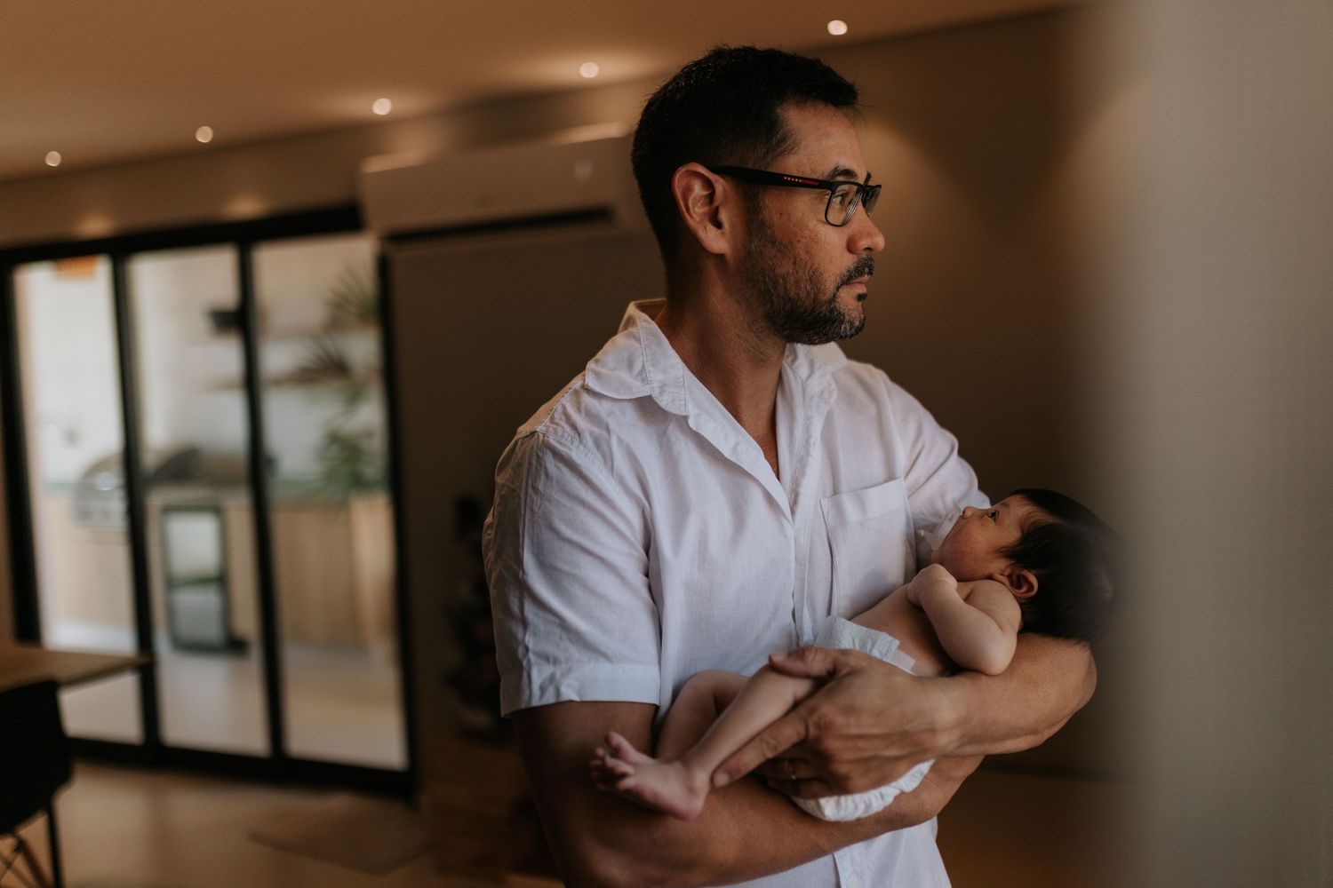 A parent in a white shirt holds a newborn baby while standing in a modern interior space.