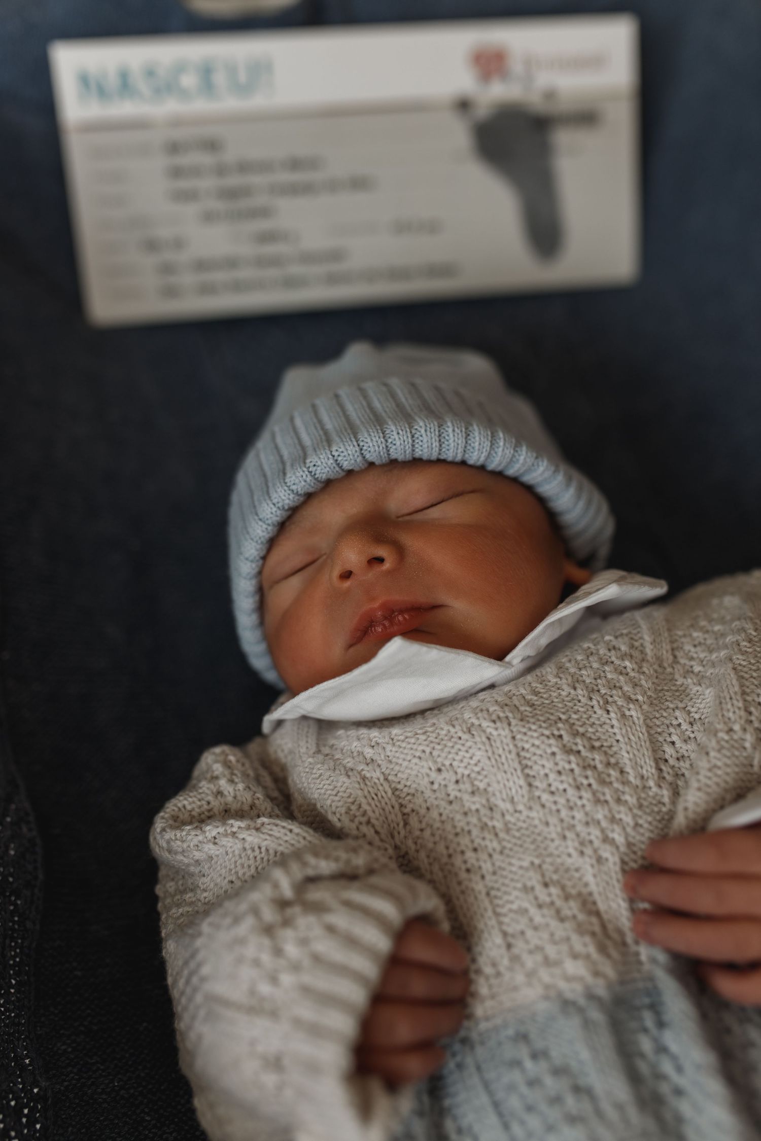 Sleeping newborn baby wearing knit hat and white sweater in hospital setting.
