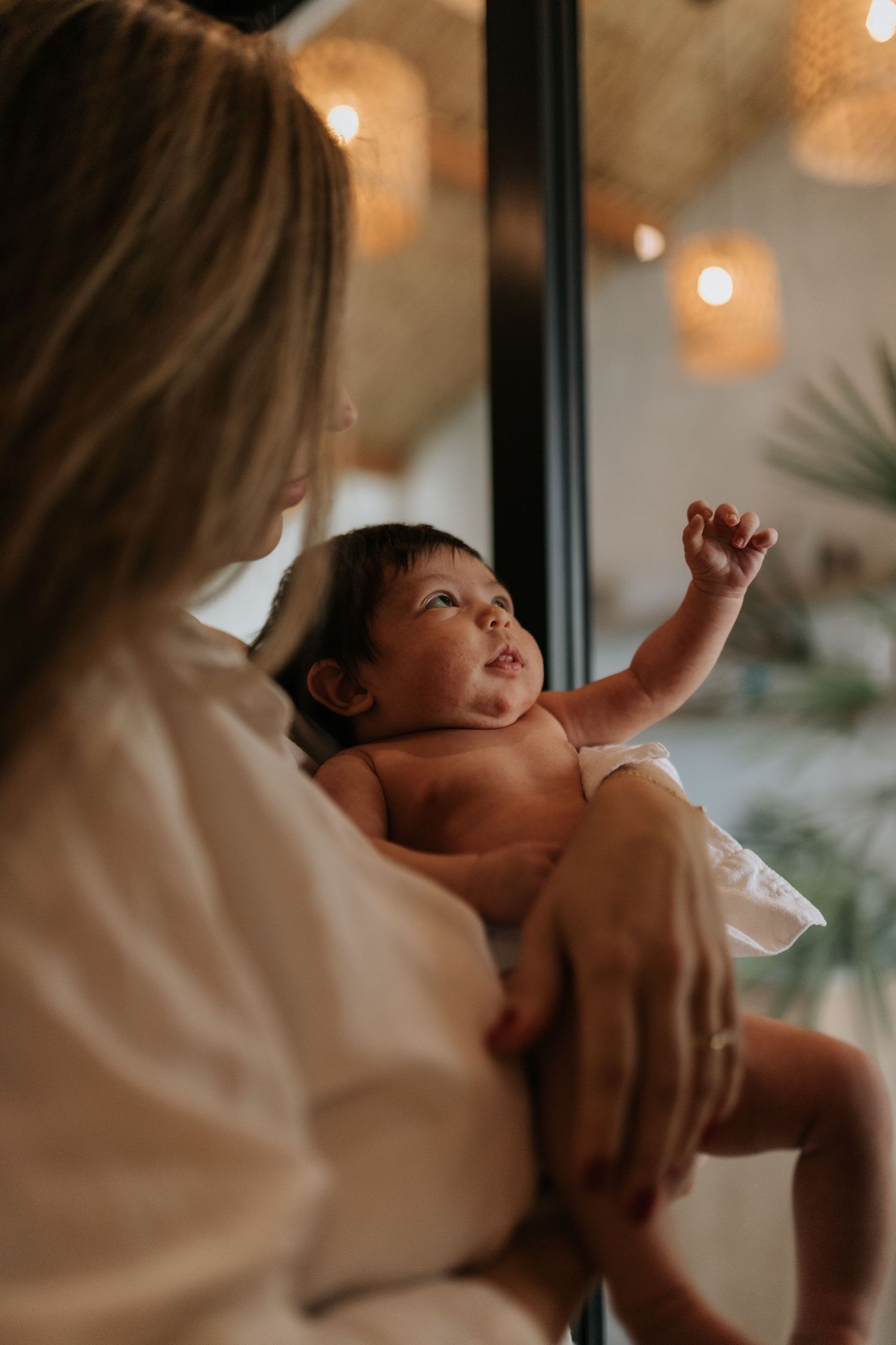 An infant reaches upward while being held against a backdrop of warm lighting and indoor plants.