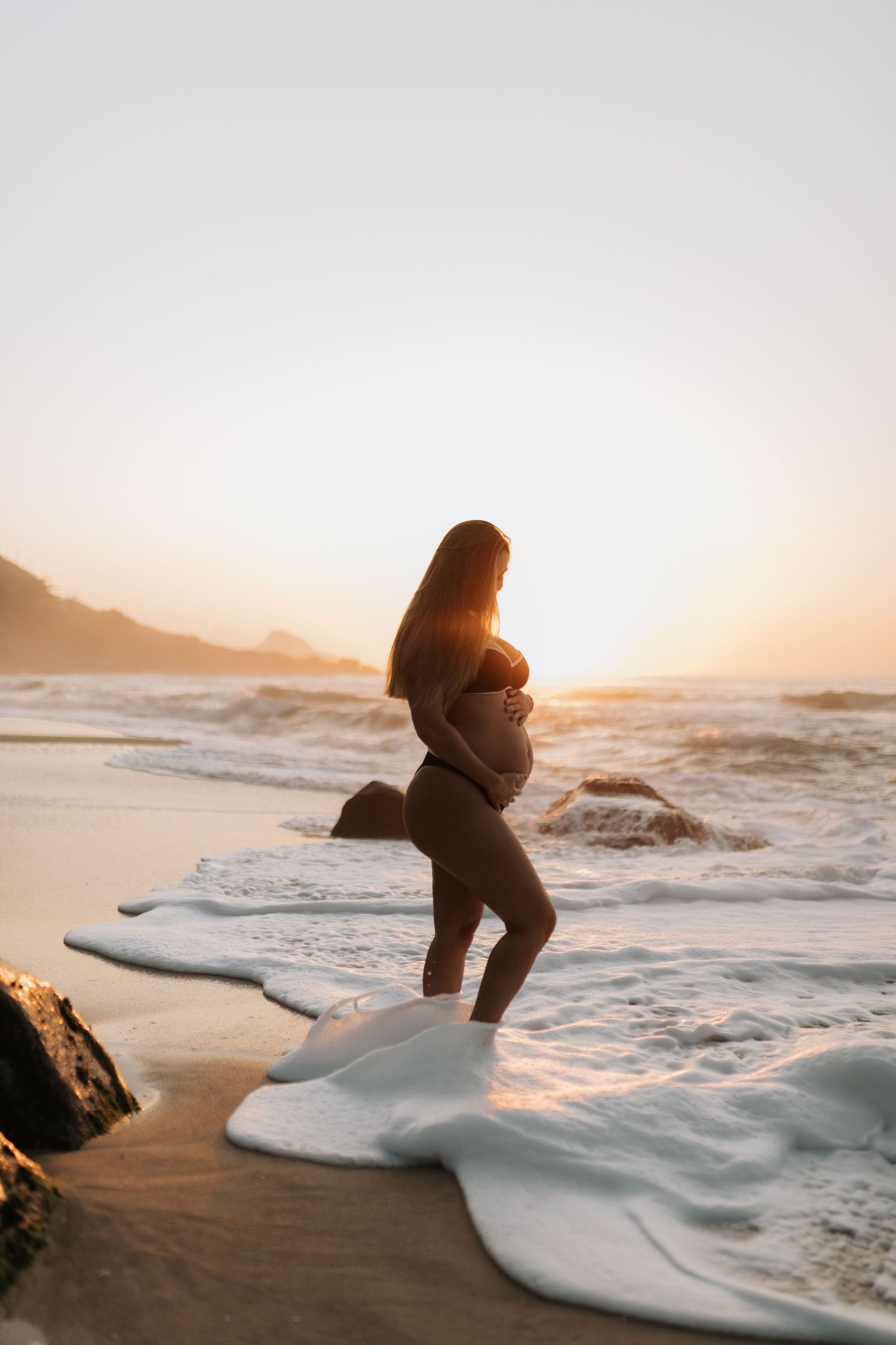 Silhouette of pregnant figure sitting on coastal rocks as waves crash during sunset.
