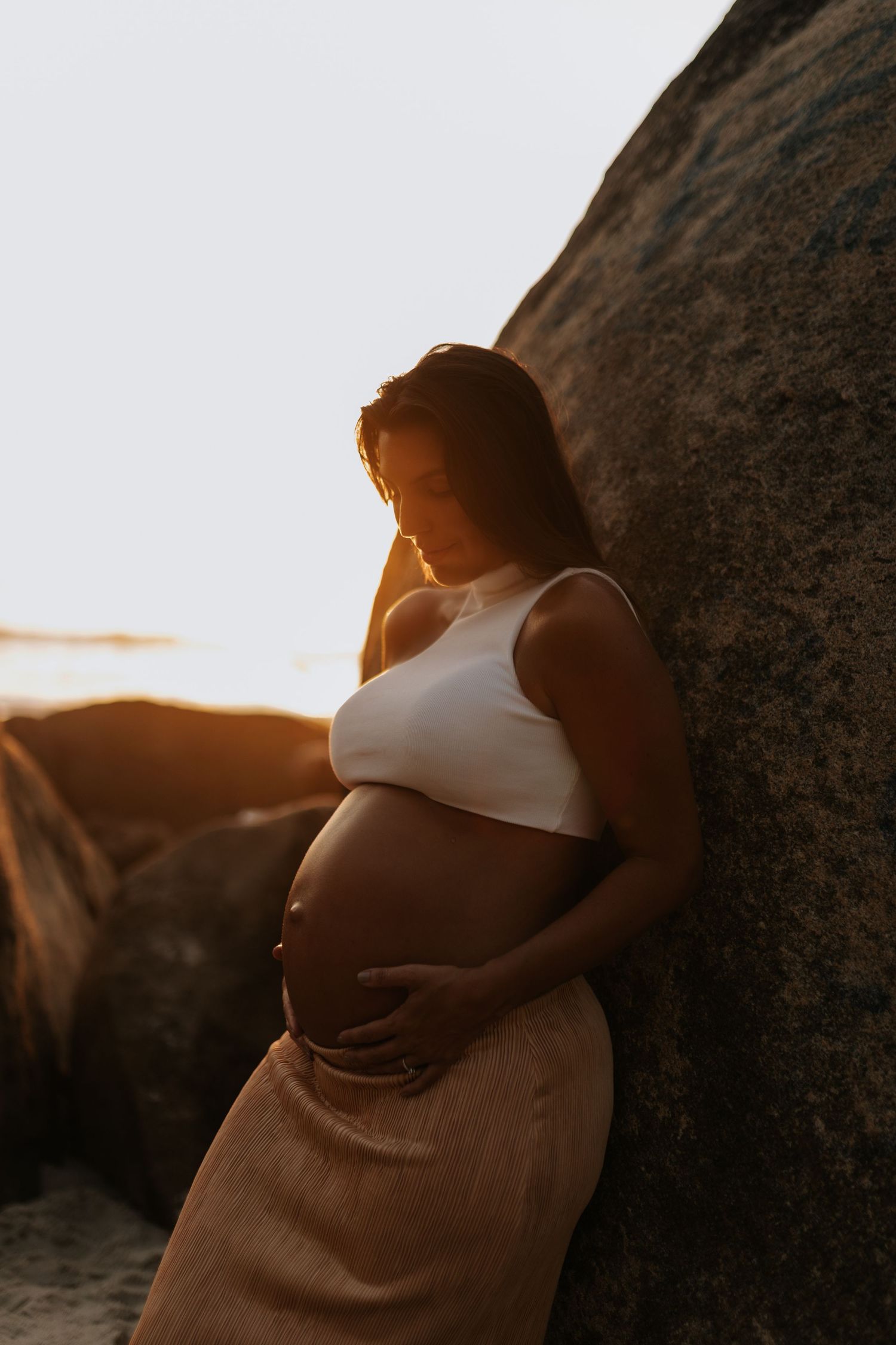 Pregnant woman in white top and brown dress poses against rocks during golden hour photoshoot.