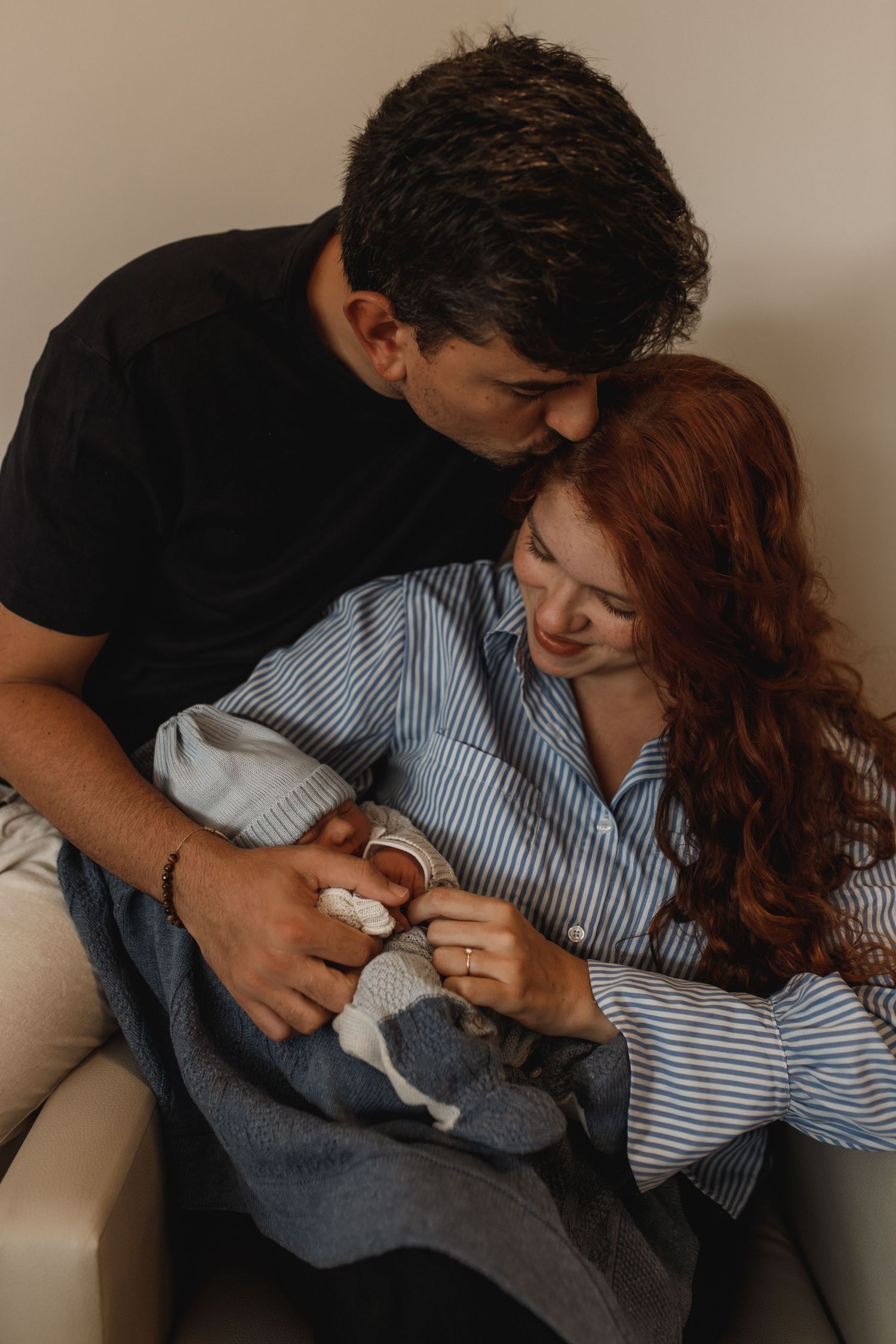 A couple shares an intimate moment while holding a small infant wrapped in blue clothing.