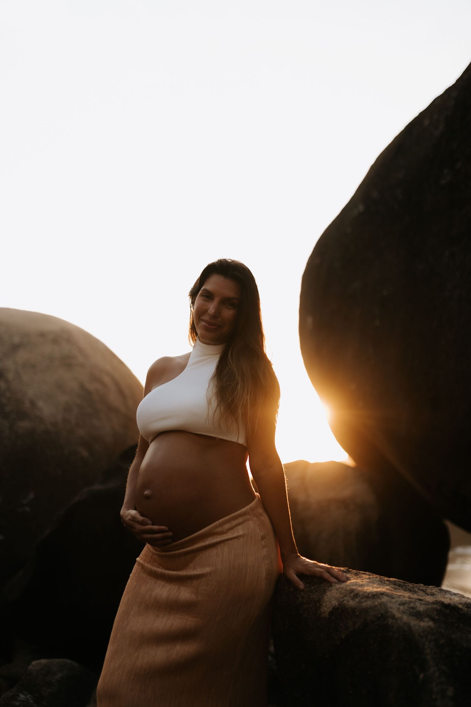 Maternity photoshoot at sunset with dramatic backlighting among coastal rocks.