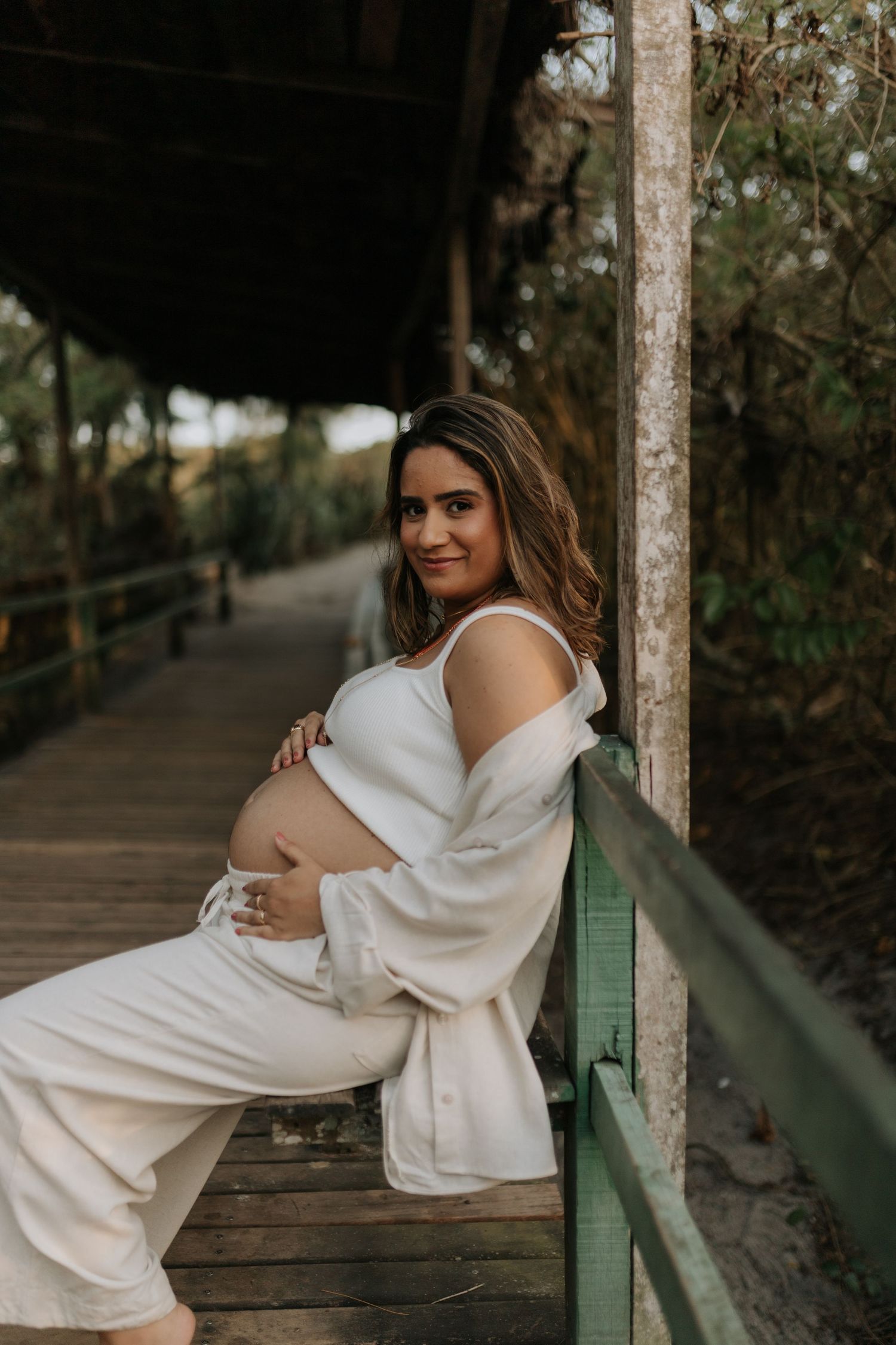 Woman in white dress posing gracefully on wooden boardwalk surrounded by trees.