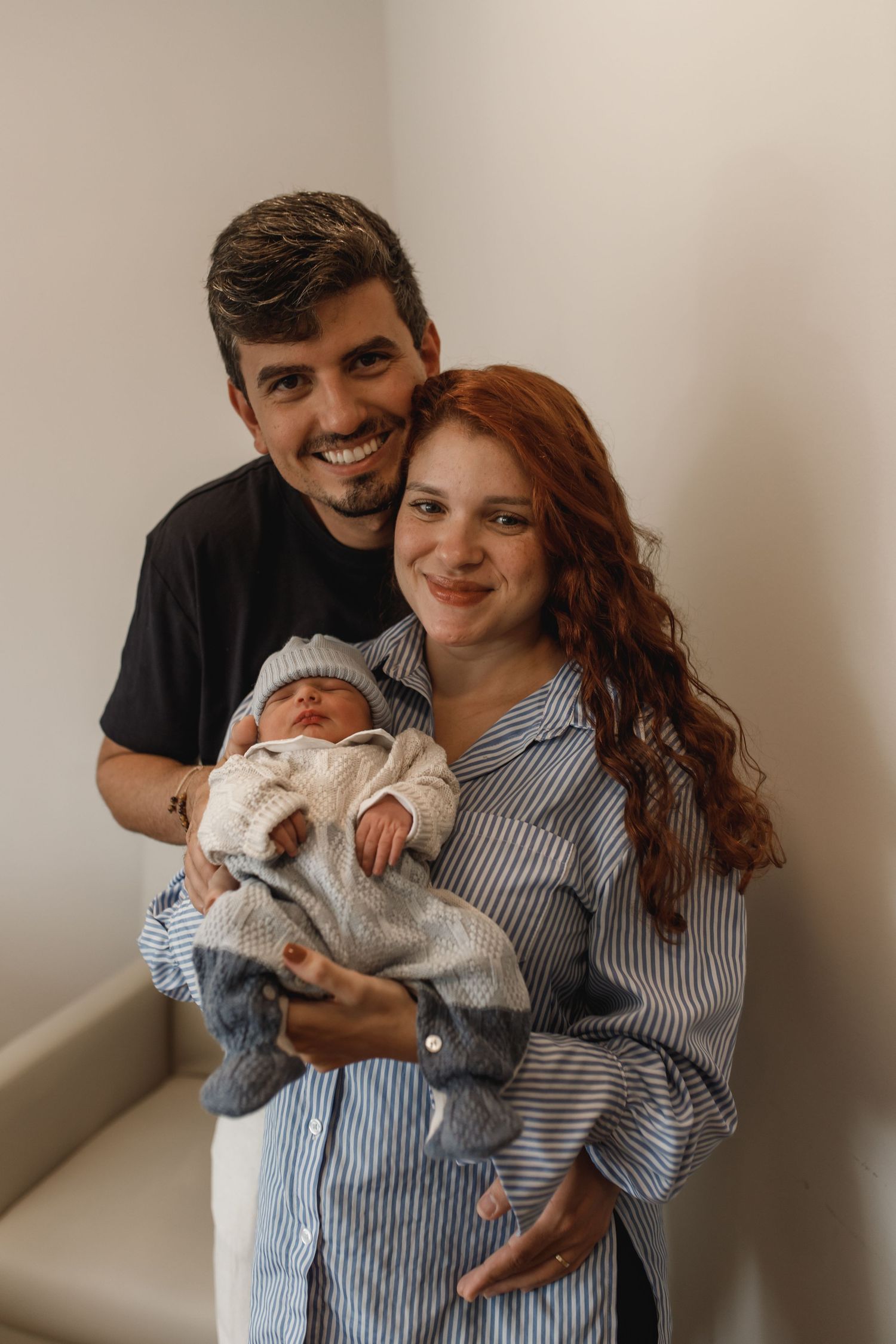 Happy parents pose with their newborn baby against a neutral wall background.