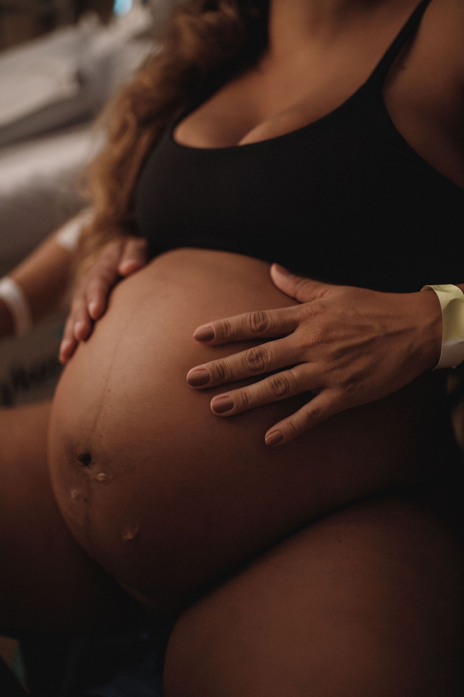Close-up of hands gently resting on pregnant belly in dim lighting.